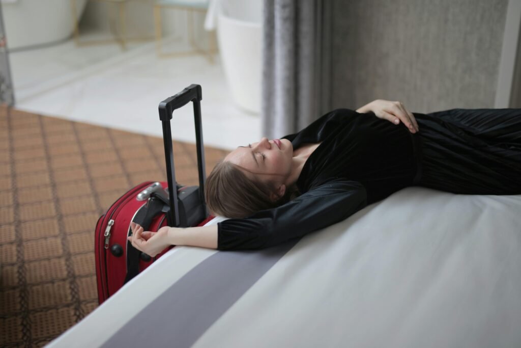 A woman resting on a hotel bed beside her suitcase, capturing travel fatigue.